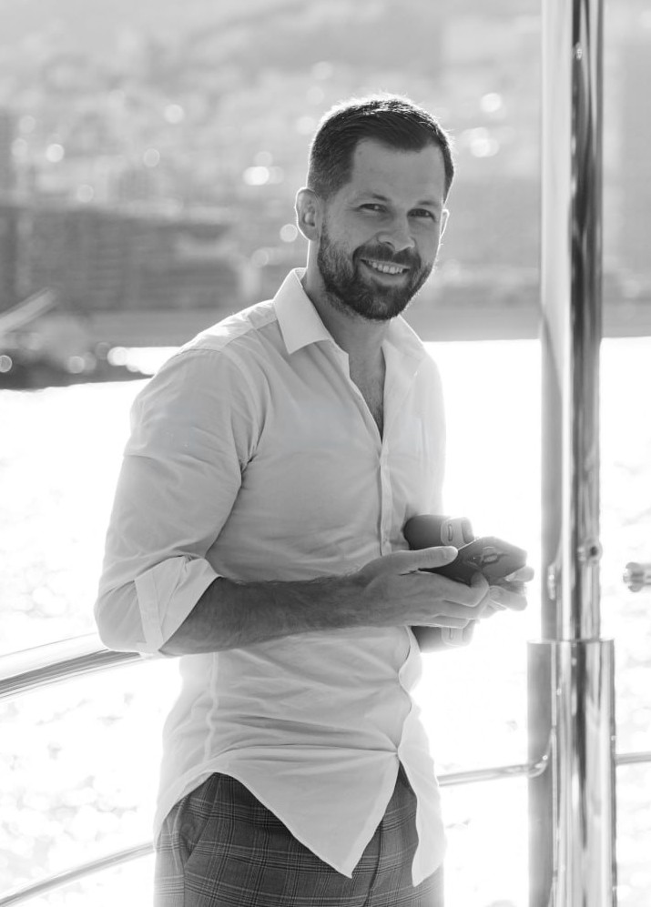 Slava smiling on the deck of a yacht, the coastline behind him